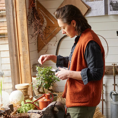 A terracotta-coloured Haws Fazeley Flow metal watering can on a work bench with a woman cutting a bonsai tree.