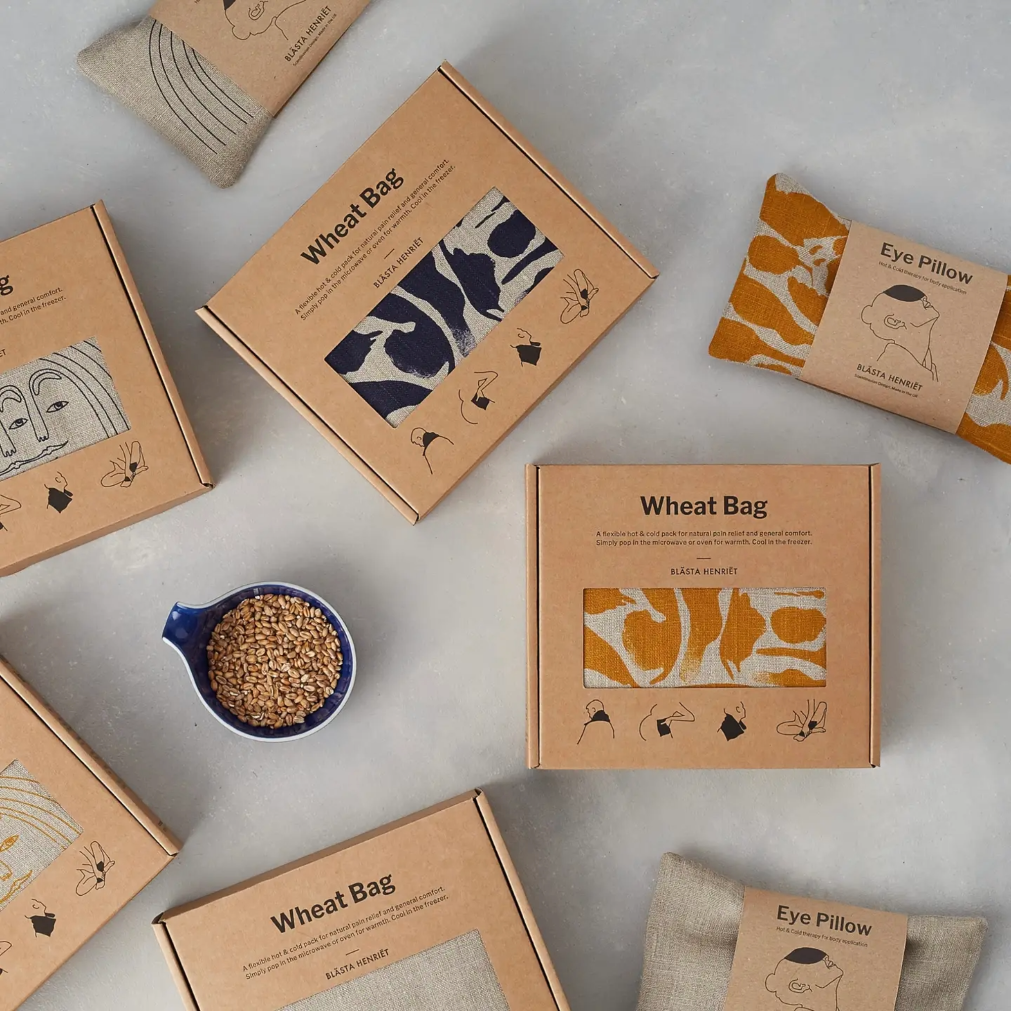 Wheat bags and eye pillows, with packaging and a cup of wheat, on a light grey surface.